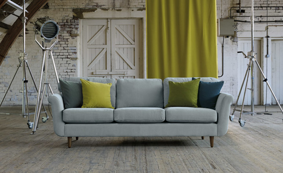 Ein hellgraues Sofa mit grünen und blaugrünen Kissen steht in der Mitte eines rustikalen Raums mit Holzböden, weißen Ziegelwänden und Studiobeleuchtungsständern und schafft eine Atmosphäre, die wie die perfekte Kulisse für einen Inneneinrichtungsservice wirkt. HDO GmbH - Ihr Handwerksdienstleister Oberschwaben - Jahnstraße 46 | 88214 Ravensburg
